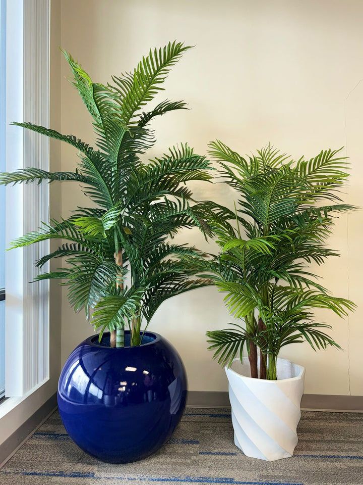 Two potted artificial tropical plants, one in a blue pot and one in a white pot, against a neutral wall.