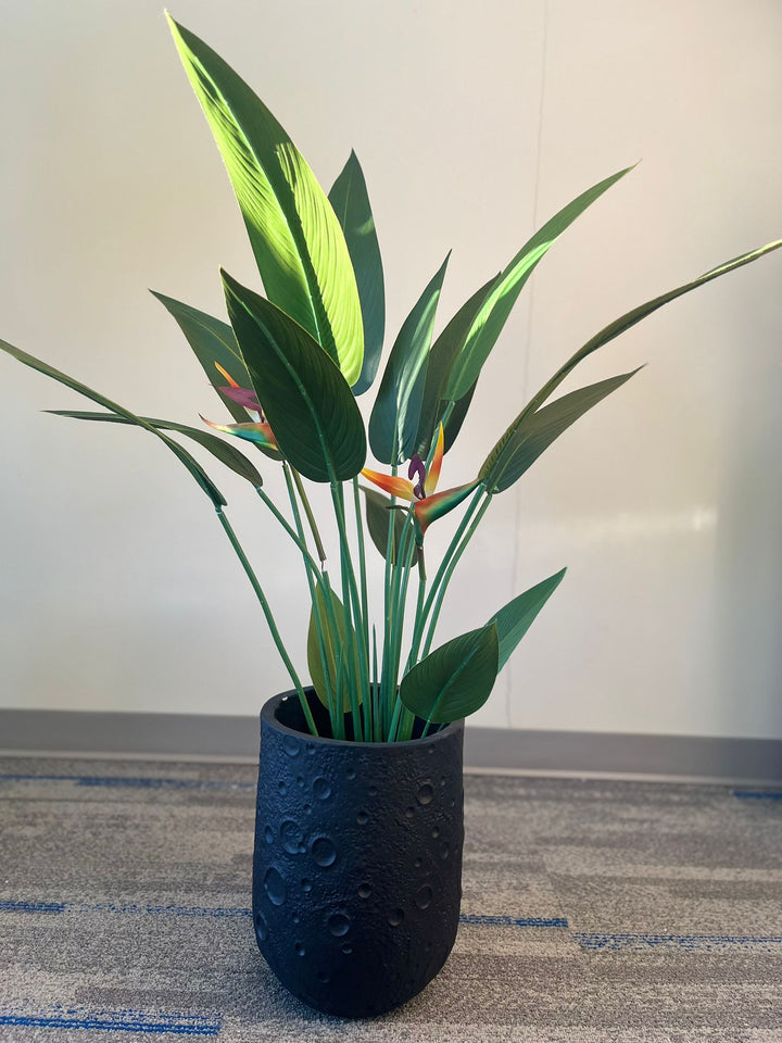Black planter with a crater-like textured design, holding an artificial bird of paradise plant with vibrant green leaves and orange flowers, placed indoors on a carpeted floor. Is this planter made from eco-friendly materials?