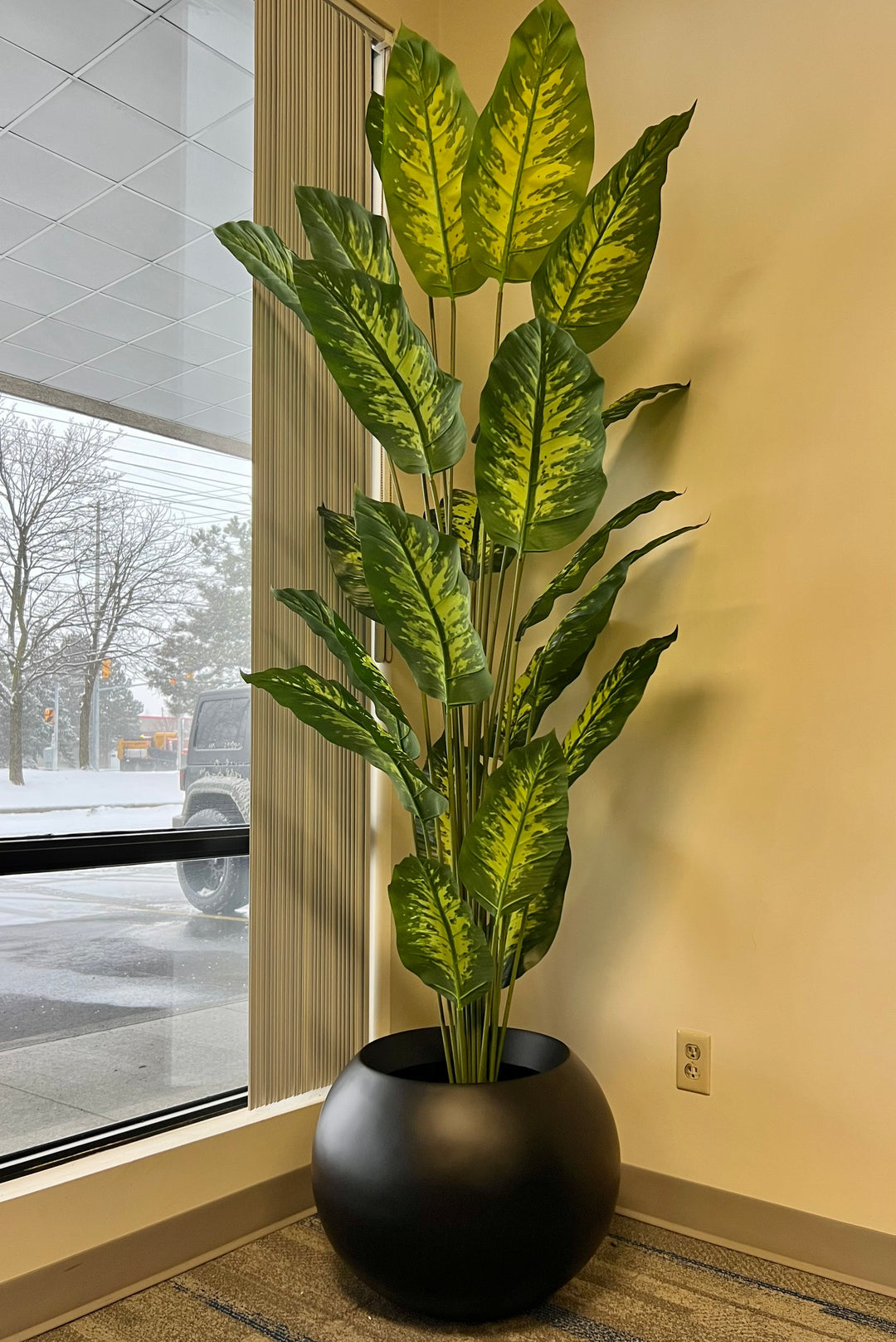 A tall, lush artificial plant with broad green leaves featuring yellow variegation is elegantly placed in a sleek, round white pot. Positioned next to a cozy white armchair, this setup adds a fresh and vibrant touch to the room's decor.