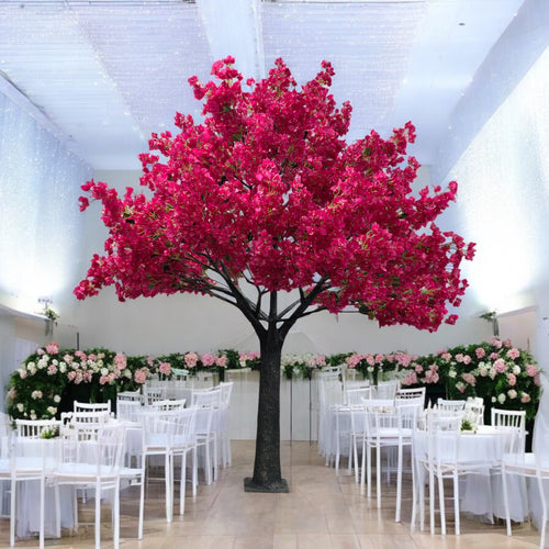 Artificial bougainvillea tree with vibrant pink flowers, prominently displayed as a centerpiece in a luxurious wedding venue setup. Surrounded by elegantly arranged white chairs and tables with floral decorations, under a beautifully lit, draped ceiling.