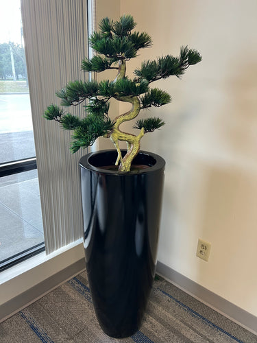 Artificial bonsai tree with a twisted trunk and lush green foliage, placed in a tall, glossy black pot, positioned near a window with vertical blinds