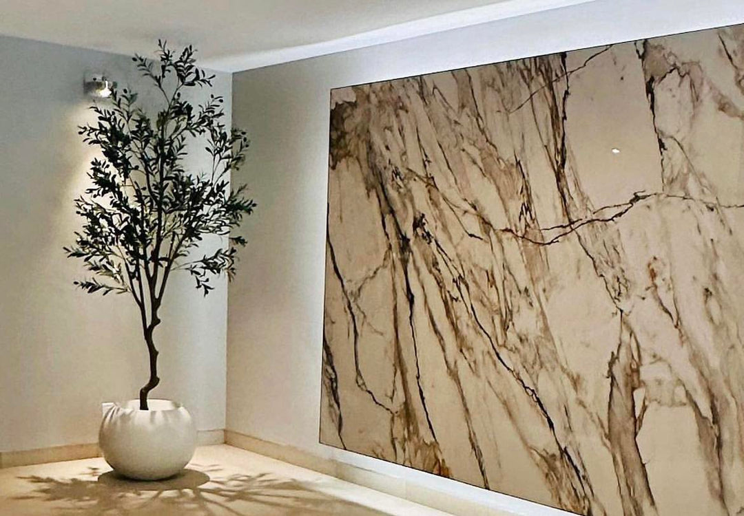 A tall artificial olive tree in a contemporary white planter is placed in a stylish indoor setting. The tree is positioned next to a large, elegant marble wall.