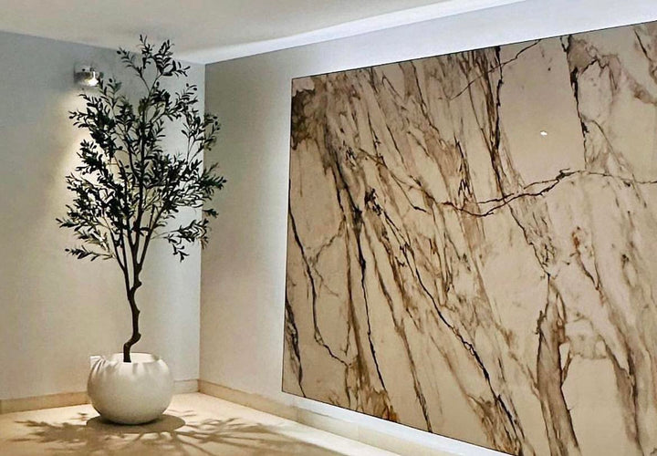 A tall artificial olive tree in a contemporary white planter is placed in a stylish indoor setting. The tree is positioned next to a large, elegant marble wall.