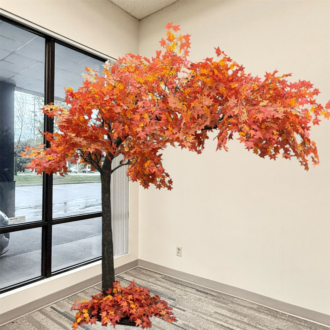 An artificial autumn maple tree with vibrant orange, red, and yellow leaves, planted in a circular base with matching foliage, set in a room corner with a window.