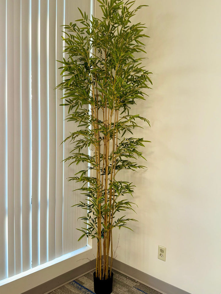 Full-height artificial bamboo tree placed in a modern interior corner, showcasing layered green leaves and natural bamboo trunks reaching toward the ceiling.