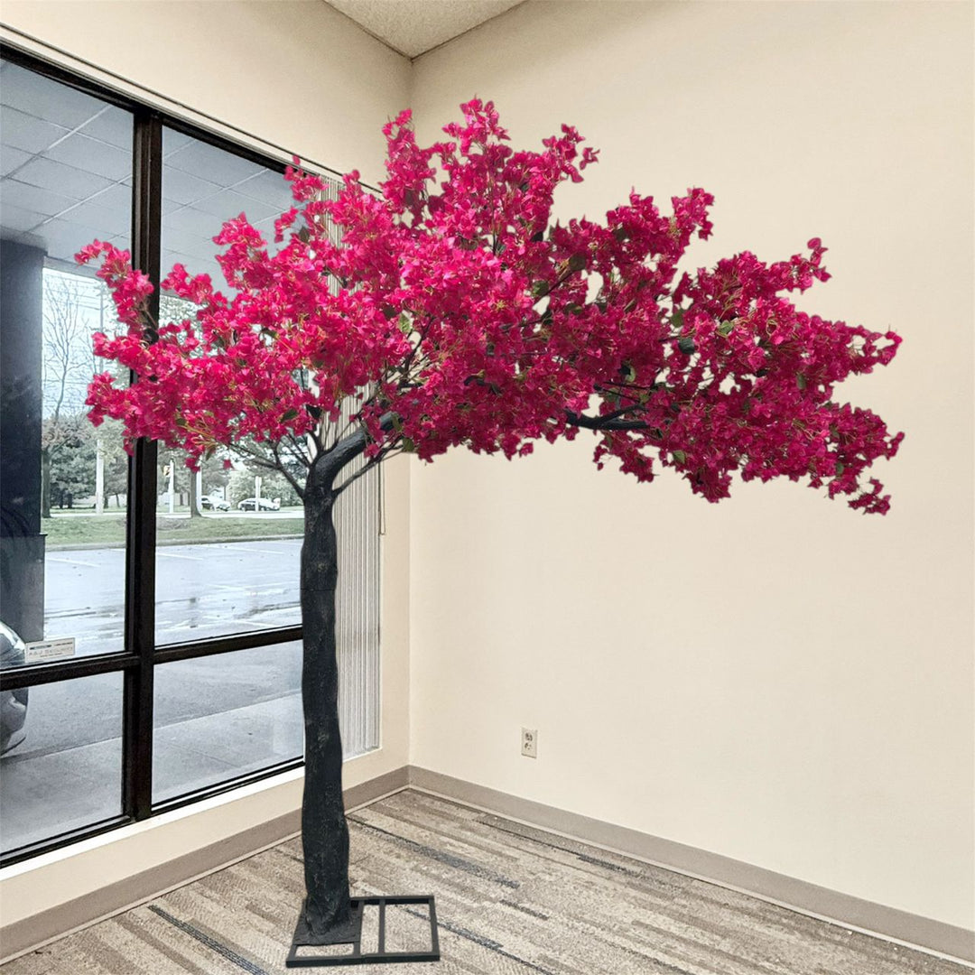 8ft artificial arch bougainvillea flowering tree with bright pink blossoms and a dark trunk, displayed indoors in a corner room with a window, beige walls, and patterned carpet.