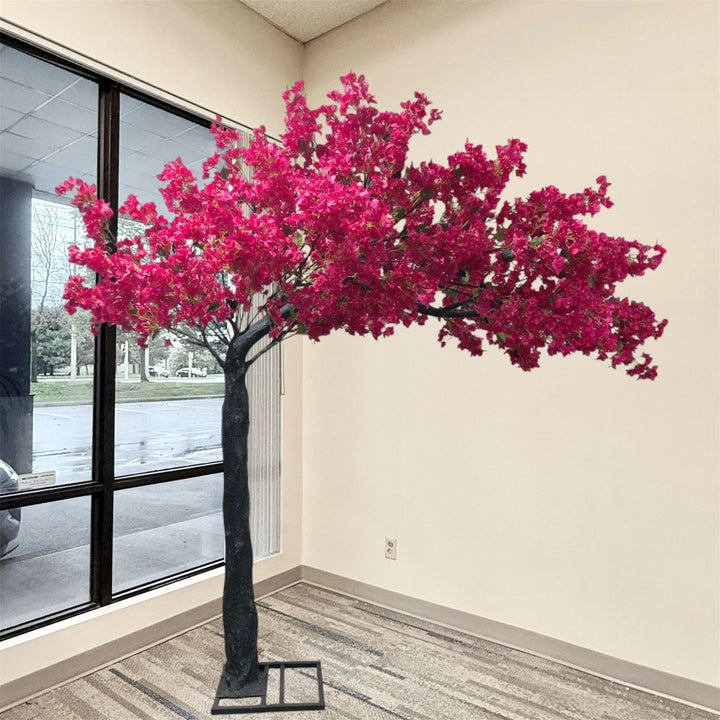 8ft artificial arch bougainvillea flowering tree with bright pink blossoms and a dark trunk, displayed indoors in a corner room with a window, beige walls, and patterned carpet.