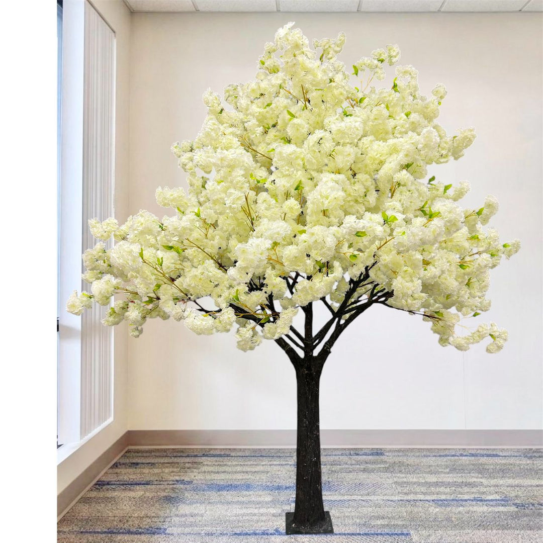 An artificial cherry blossom tree with dense cream white flowers with green leaves and a dark trunk, displayed indoors on a patterned carpet against a plain wall.