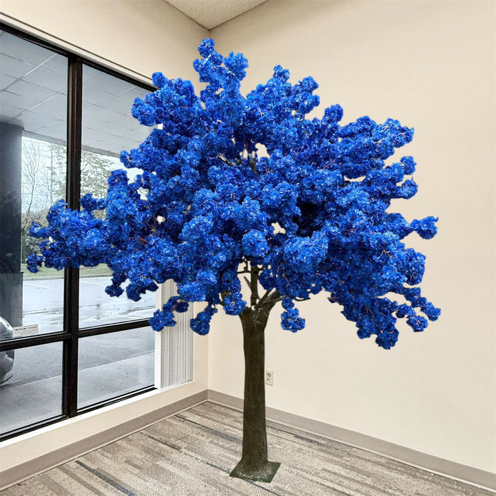 8ft artificial blue flowering tree with a dark brown trunk stands indoors near a window in a room with beige walls and a striped floor.