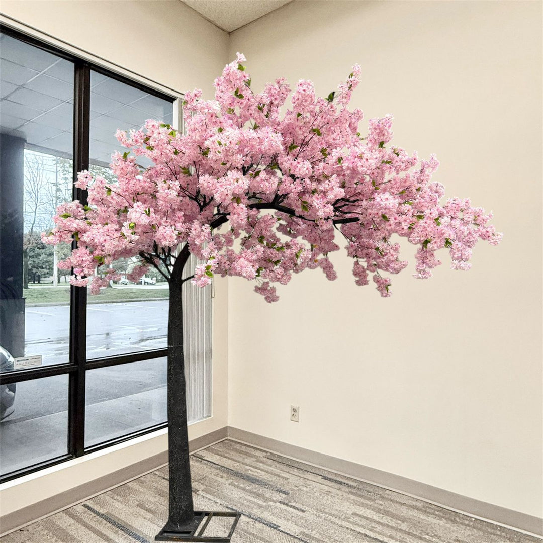 An artificial purple arch cherry blossom tree with dense purple blossoms and a dark trunk, displayed indoors against a beige wall near a large window.