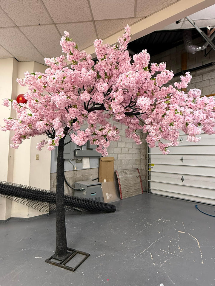 An artificial cherry blossom tree with dense purple blossoms and a dark trunk, standing on a metal base in a garage or workshop setting.