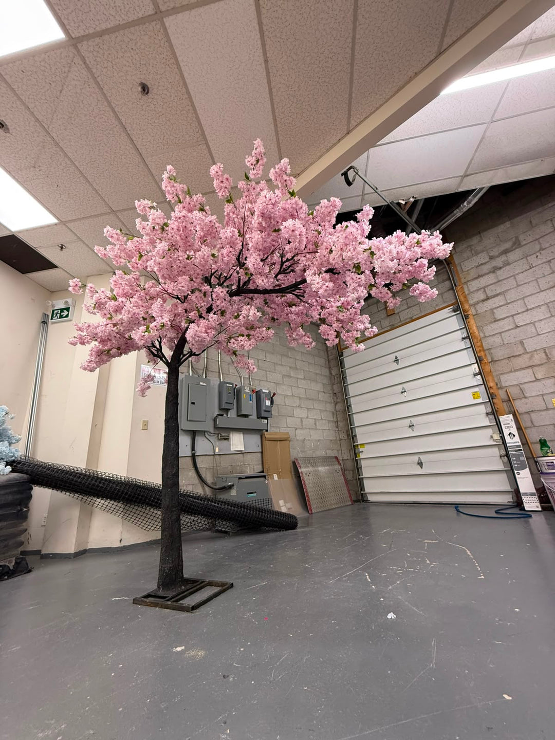 An artificial cherry blossom tree with purple blossoms and a dark trunk, standing on a gray floor inside a warehouse or garage with a white door and ceiling tiles in the background.