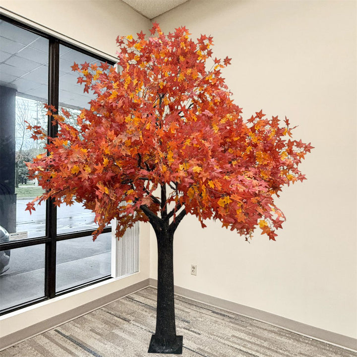 An artificial autumn maple tree with vibrant red, orange, and yellow leaves on a black trunk, standing indoors near a window.