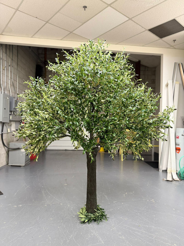Artificial olive tree in an indoor warehouse setting with a tiled ceiling.