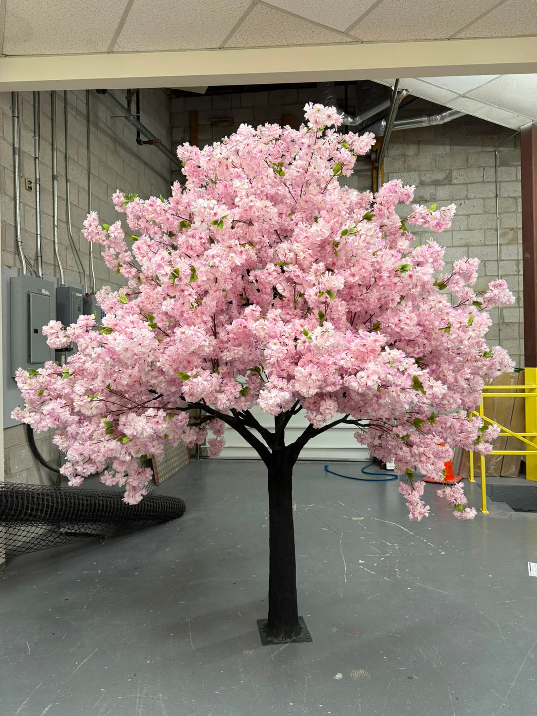 An artificial cherry blossom tree with dense purple blossoms and a dark trunk, standing on a gray floor in an indoor warehouse or showroom setting.