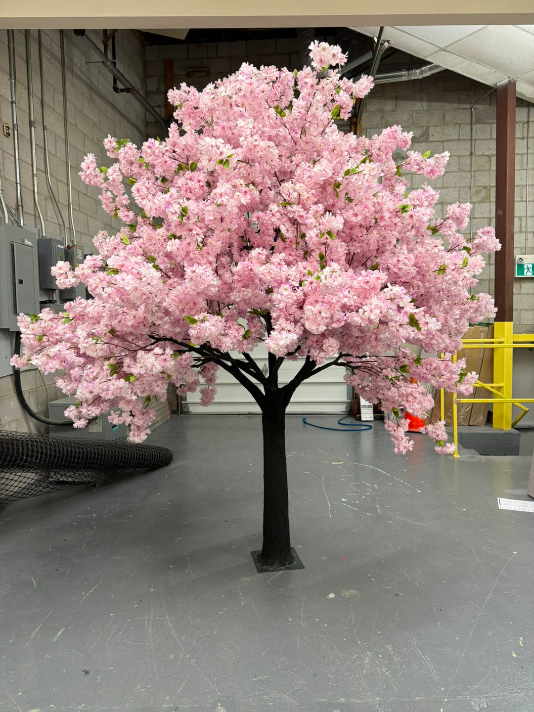 An artificial cherry blossom tree with dense purple blossoms with green leaves and a dark trunk, standing on a gray floor inside a warehouse or studio space.