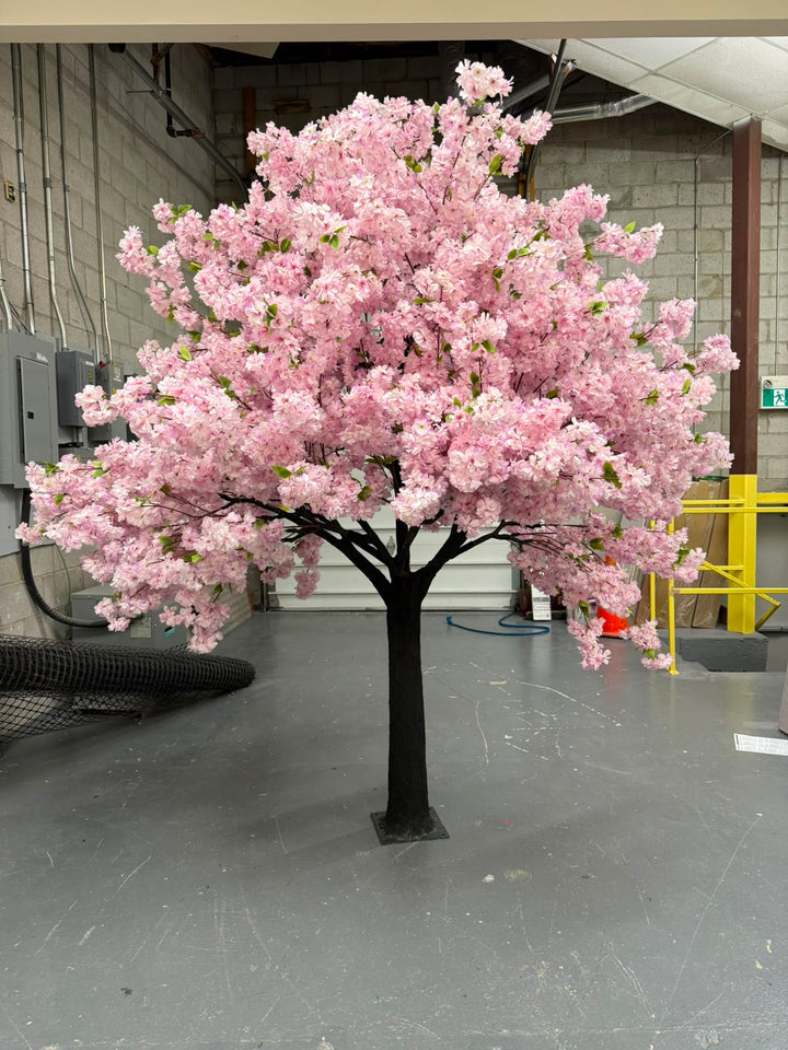 An artificial cherry blossom tree with dense purple blossoms with green leaves and a dark trunk, standing on a gray floor inside a warehouse or studio space.
