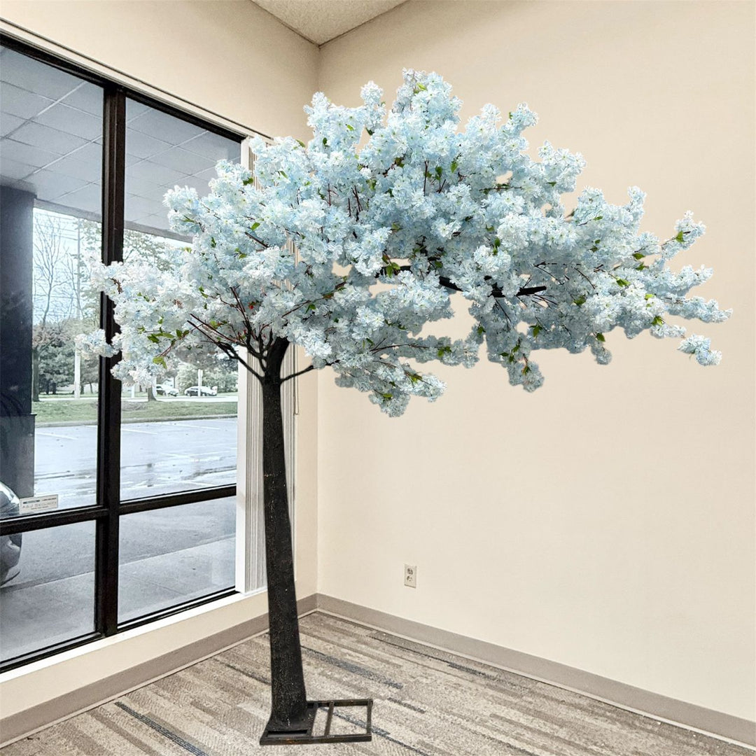 8ft soft blue arch artificial cherry blossom tree with pale blue flowers and a dark trunk, displayed indoors in a room corner with a window, beige walls, and patterned carpet.