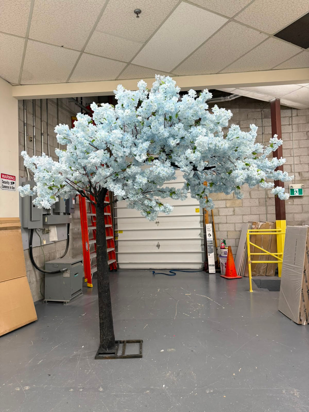 An artificial flowering tree with soft blue blossoms and a dark trunk, standing in a warehouse or showroom with a gray floor, white garage door, and storage items in the background.