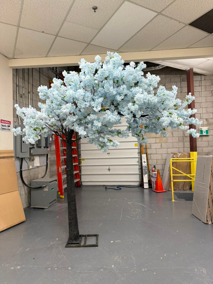 An artificial flowering tree with soft blue blossoms and a dark trunk, standing in a warehouse or showroom with a gray floor, white garage door, and storage items in the background.