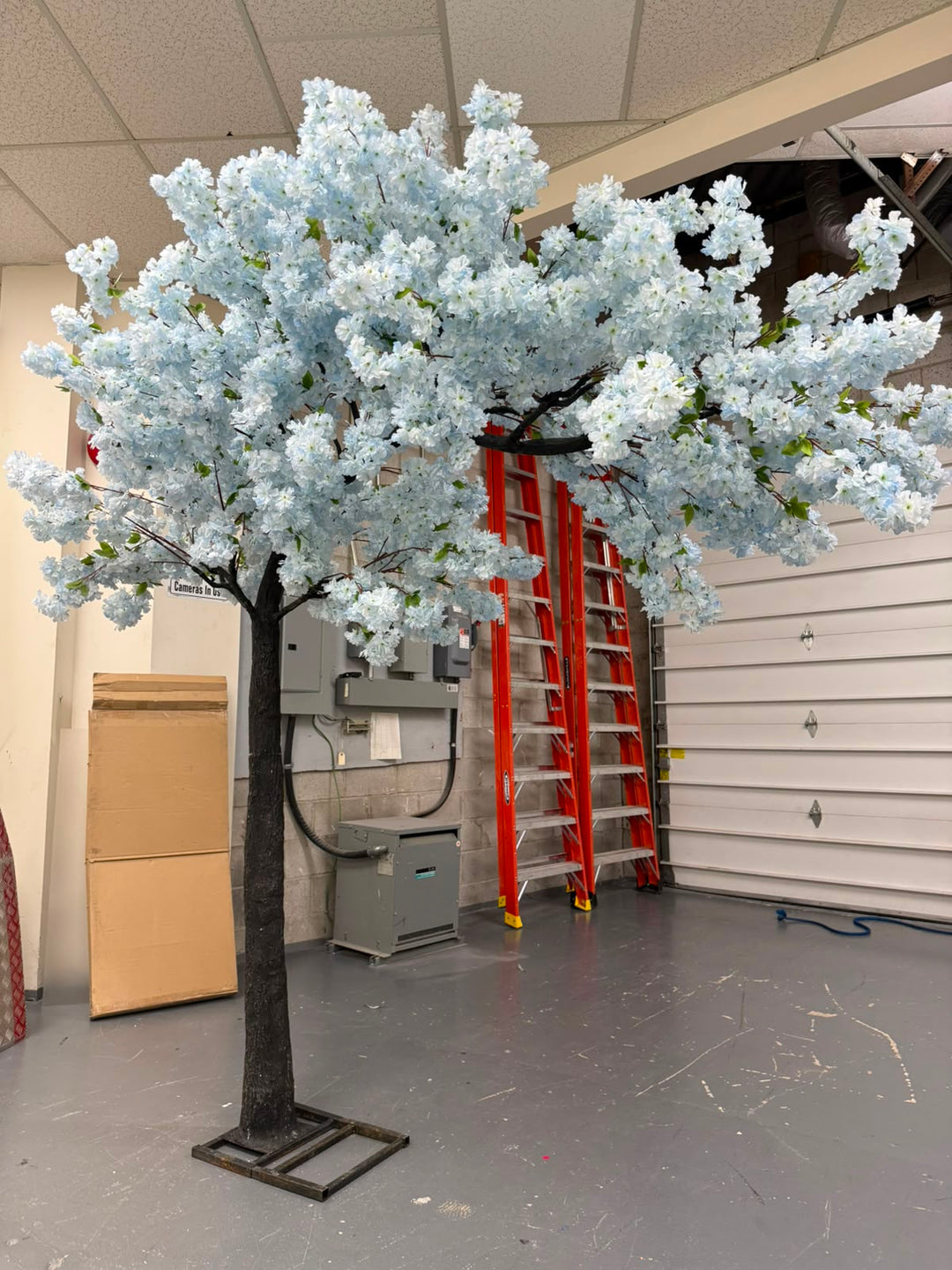An artificial cherry blossom tree with dense pale blue flowers with green leaves, set in a warehouse or garage environment with a red ladder and gray floor visible in the background.