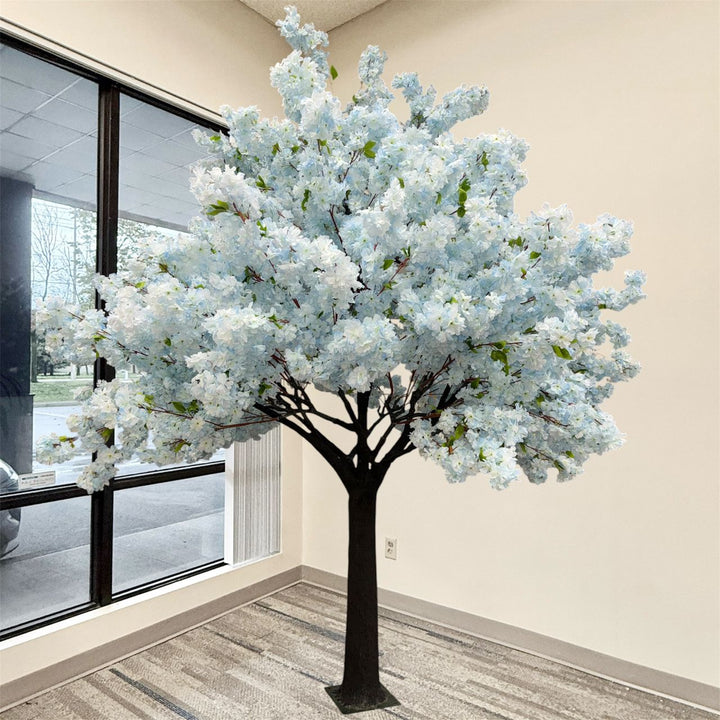 An artificial soft blue cherry blossom flowering tree with dense pale blue  blossoms, green leaves, and a dark brown trunk, displayed indoors near a window in a beige room.