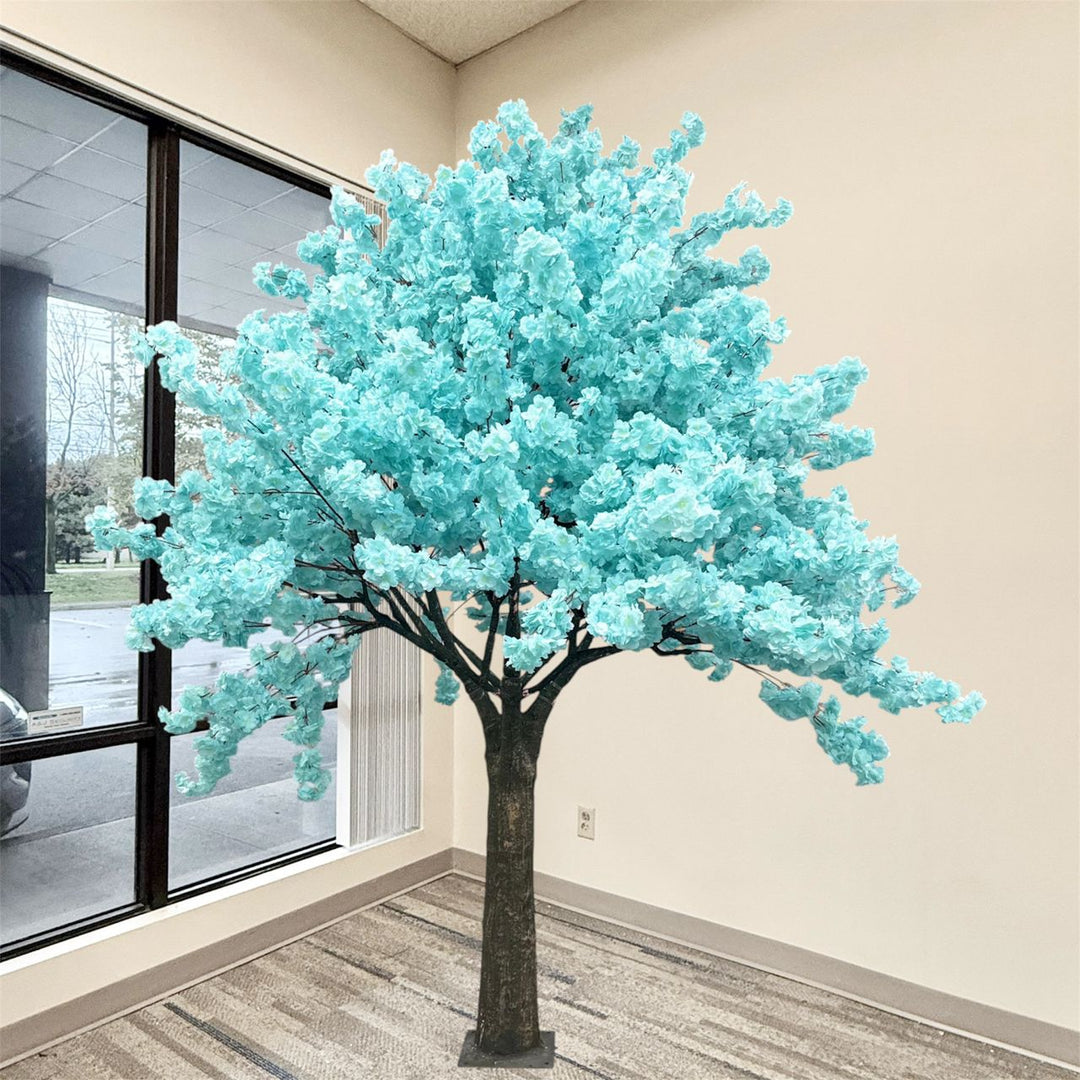 Soft blue artificial cherry blossom tree with bright turquoise-blue flowers and a dark brown trunk, displayed indoors in a room with beige walls, a window, and a patterned floor.