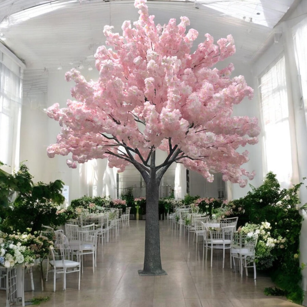 A large artificial pink cherry blossom tree placed in an elegant event space surrounded by white chairs and floral decorations.