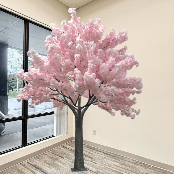 8ft  artificial cherry blossom tree with dense light pink blossoms and a dark gray trunk, placed indoors near a window in a room with beige walls and patterned carpet.