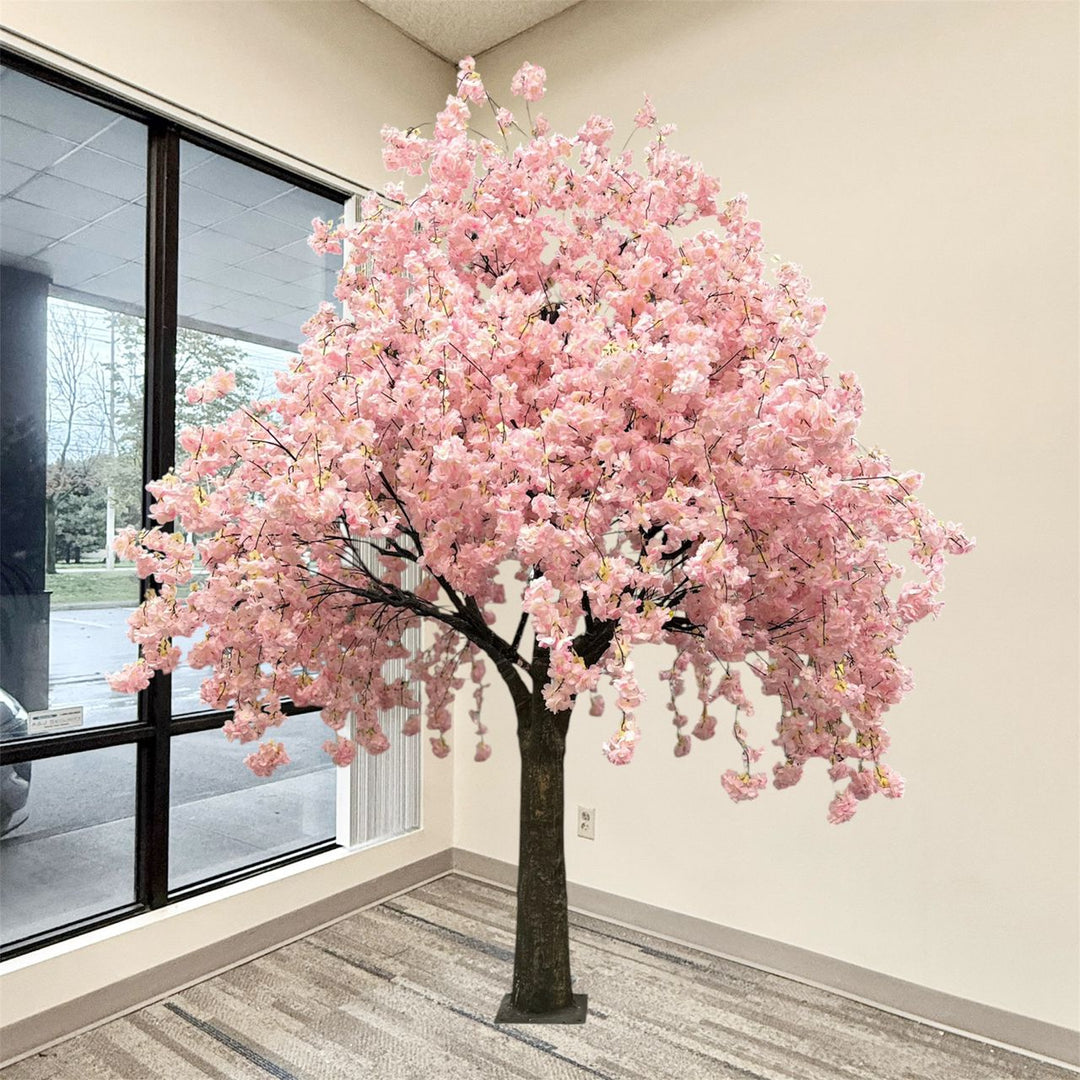 An artificial weeping cherry blossom tree with dense pink blossoms and a dark trunk, displayed indoors near a large window with beige walls and a patterned carpet.