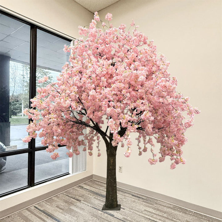 An artificial weeping cherry blossom tree with dense pink blossoms and a dark trunk, displayed indoors near a large window with beige walls and a patterned carpet.