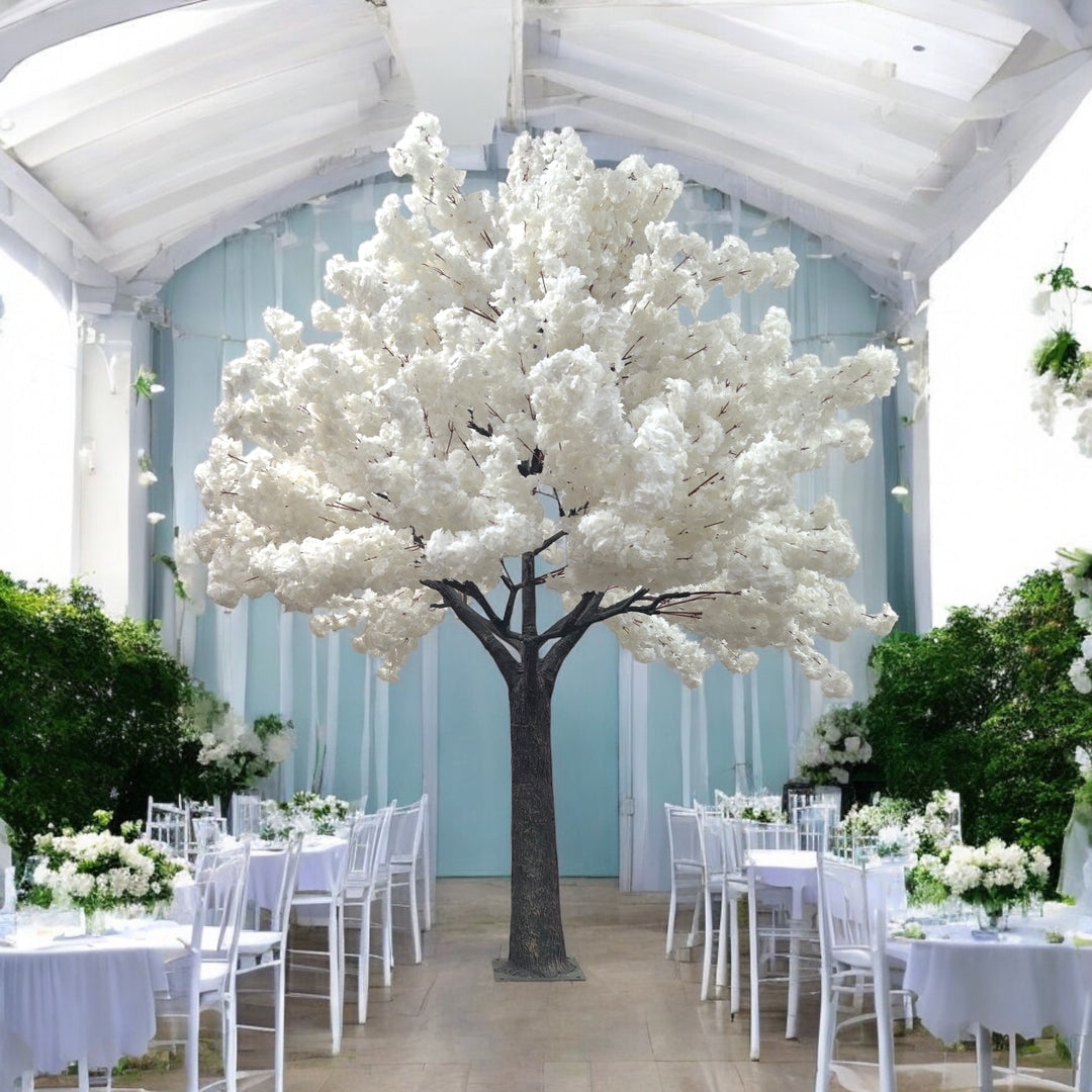 Elegant white artificial cherry blossom tree standing tall in a beautifully decorated wedding venue with white and pastel floral arrangements.