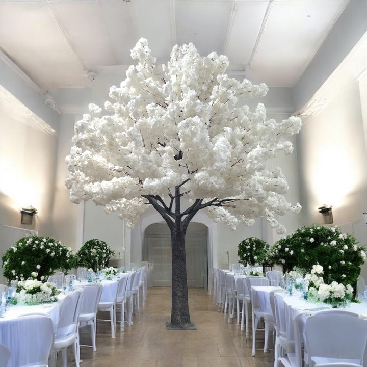 Elegant white artificial cherry blossom tree standing tall in a beautifully decorated wedding venue with white and pastel floral arrangements.