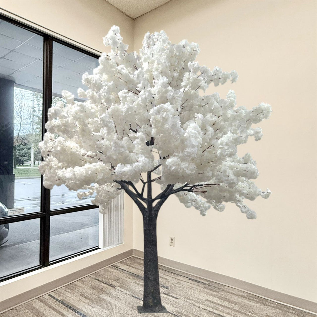 An artificial flowering white cherry blossom  tree with dense white blossoms and a dark brown trunk, standing indoors near a large window with beige walls and a patterned carpet.