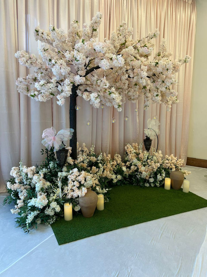 Decorative white pink arch cherry blossom floral arrangement with cherry blossom trees and candles on a green mat against a beige curtain.