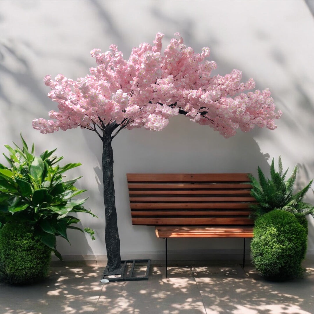 Elegant artificial light pink cherry blossom tree placed next to a wooden bench and lush greenery, creating a serene outdoor-inspired setting. Can this cherry blossom tree be rented for outdoor-themed events?