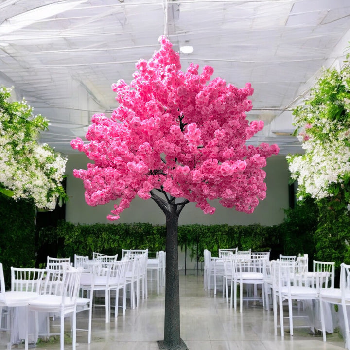 A stunning artificial pink cherry blossom tree is prominently displayed in the center of an elegant event venue. The room features white chairs and tables adorned with pastel floral arrangements, set against a backdrop of lush greenery and a high, translucent ceiling that allows soft natural light to fill the space.