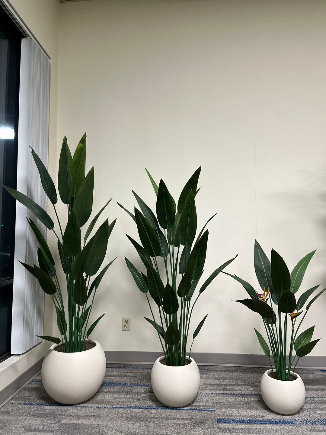 Three ivory round planters with a smooth matte finish, each holding tall artificial plants, including bird of paradise and leafy green plants, arranged indoors against a neutral background. Are these planters suitable for both indoor and outdoor use?