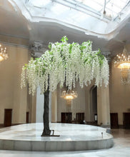 Charger l&#39;image dans la galerie, A tall, elegant artificial Wisteria tree in white, featuring dense foliage and hanging blooms, placed inside a storage area with boxes in the background.
