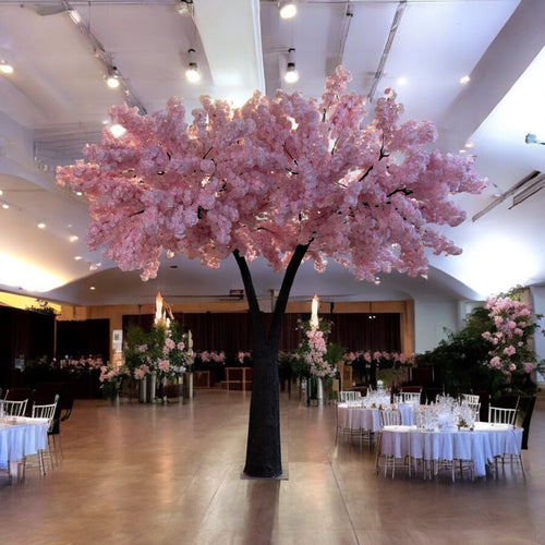 Artificial tree adorned with soft pink blossoms, beautifully displayed as a centerpiece in an elegantly decorated event hall with round dining tables and floral arrangements. Can these rental trees enhance the ambiance of formal events like weddings and banquets?