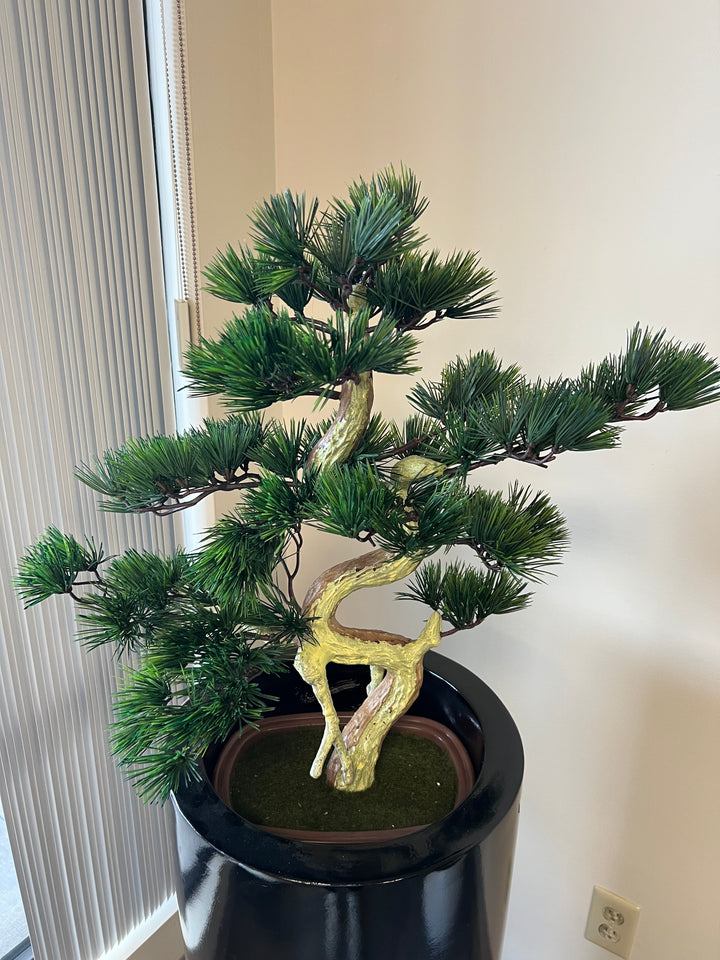 Artificial bonsai tree with a twisted trunk and dense, needle-like green foliage, displayed in a shiny black planter, situated near a window with vertical blinds