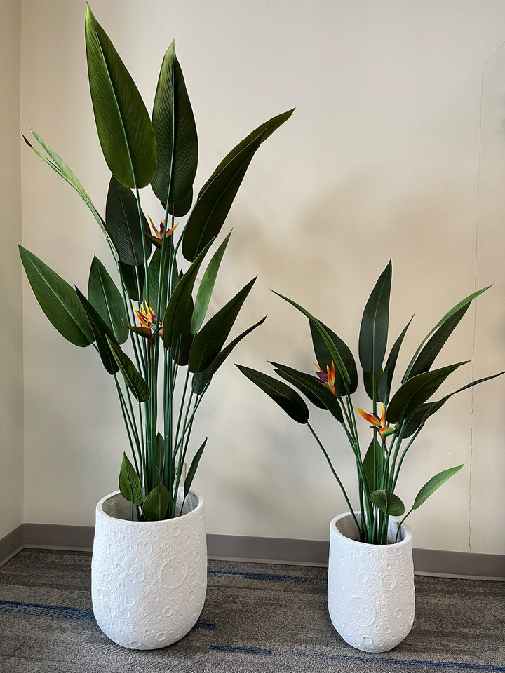 White planters with a unique textured design resembling craters, each holding artificial bird of paradise plants with vibrant green leaves and orange flowers, placed indoors on a carpeted floor. Are these planters weather-resistant for outdoor use?