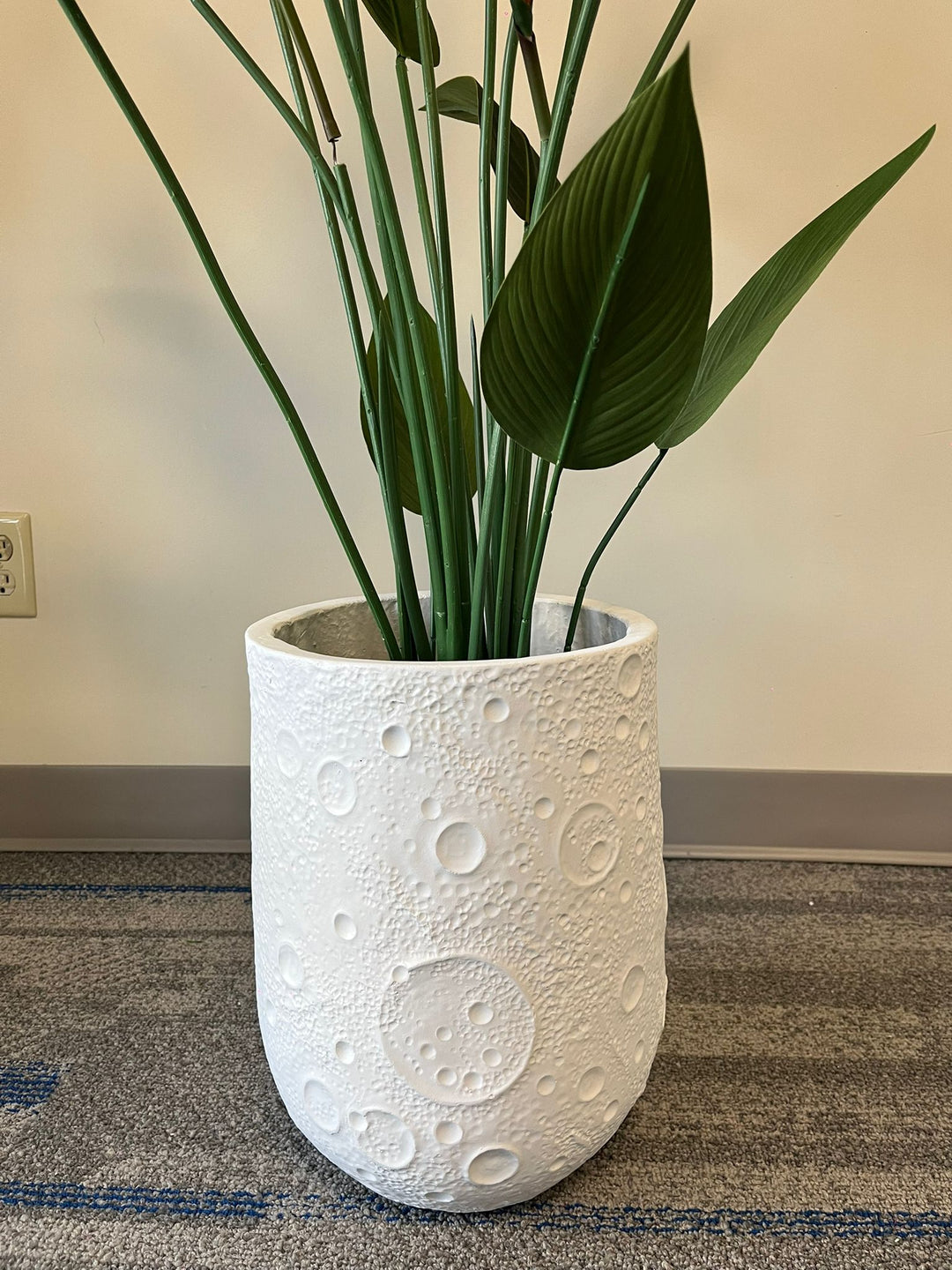 White planter with a crater-like textured design, holding an artificial bird of paradise plant with vibrant green leaves and orange flowers, placed indoors on a carpeted floor. Is this planter made from eco-friendly materials?