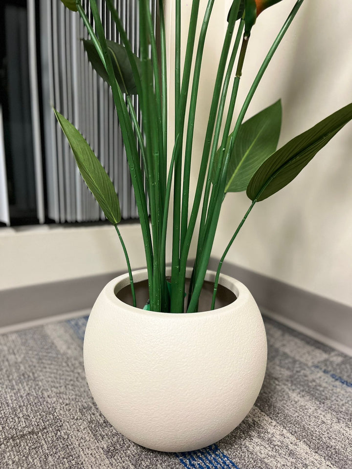 Close-up of an ivory round planter with a smooth matte finish, holding an artificial palm plant with vibrant green leaves, placed indoors near a window. Does this planter have a built-in drainage system?
