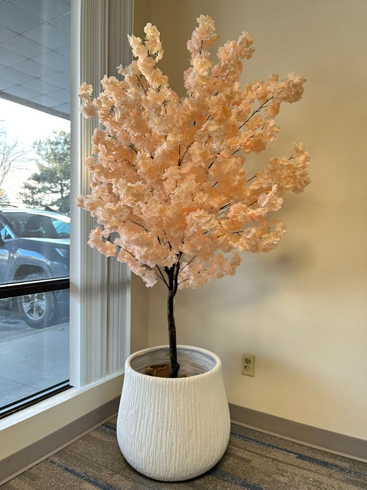 White textured planter with a ribbed design, holding a tall artificial peach blossom tree with delicate pink flowers, placed indoors near a window. Is this planter stable for taller artificial trees?