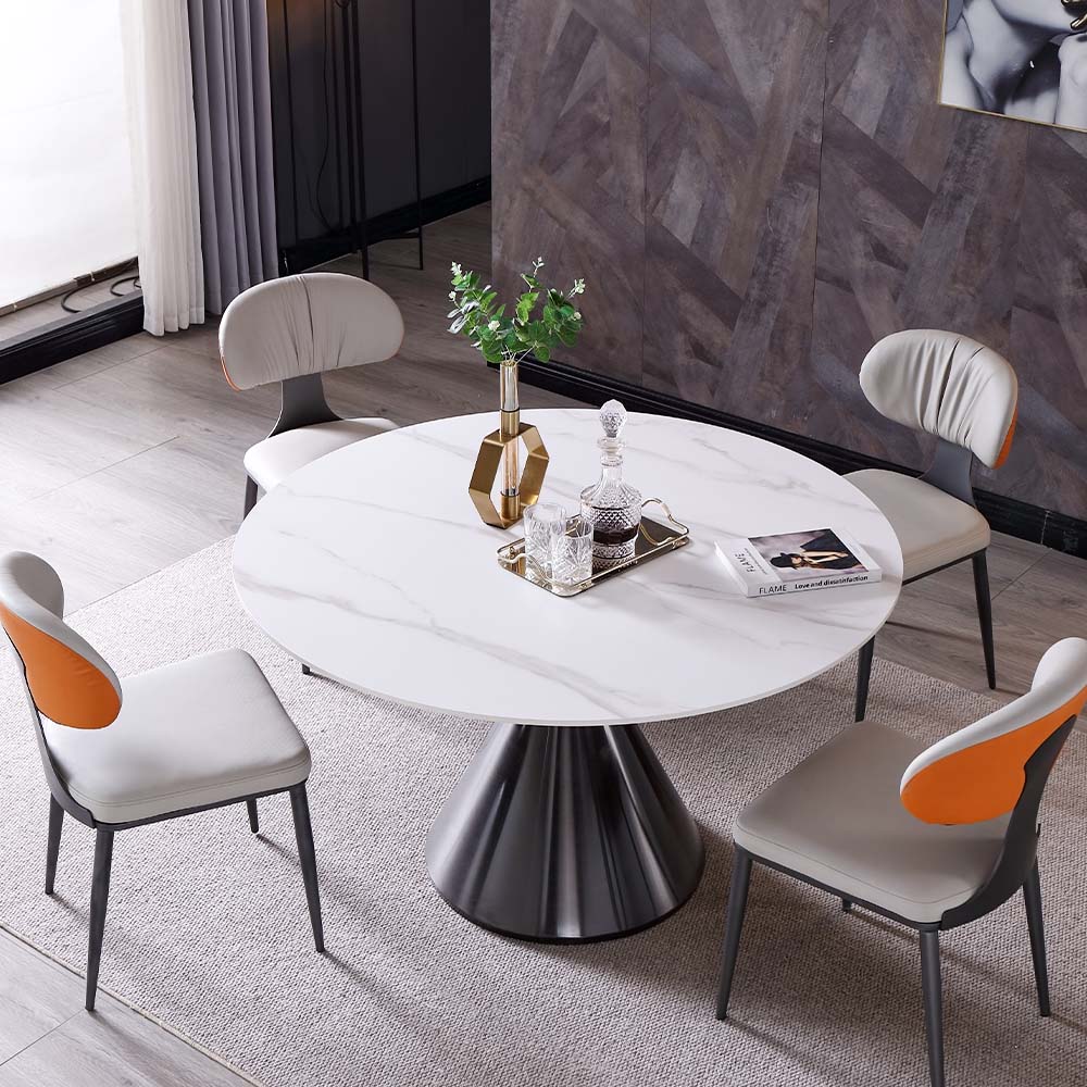 Alt text: Modern round dining table with white top and black tulip base, surrounded by beige chairs with orange accent cushions, decorative vase with greenery, crystal decanter set, magazine, and gray area rug in stylish dining space with dark curtains and marble accent wall.