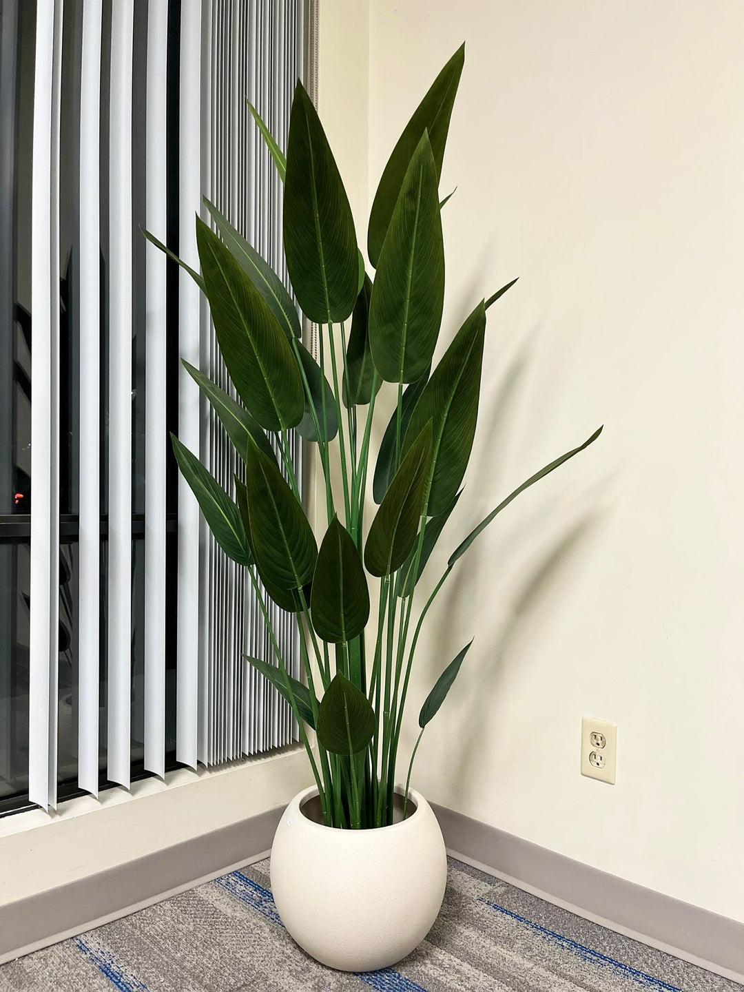 Close-up of an ivory round planter with a smooth matte finish, holding an artificial palm plant with vibrant green leaves, placed indoors near a window. Does this planter have a built-in drainage system?