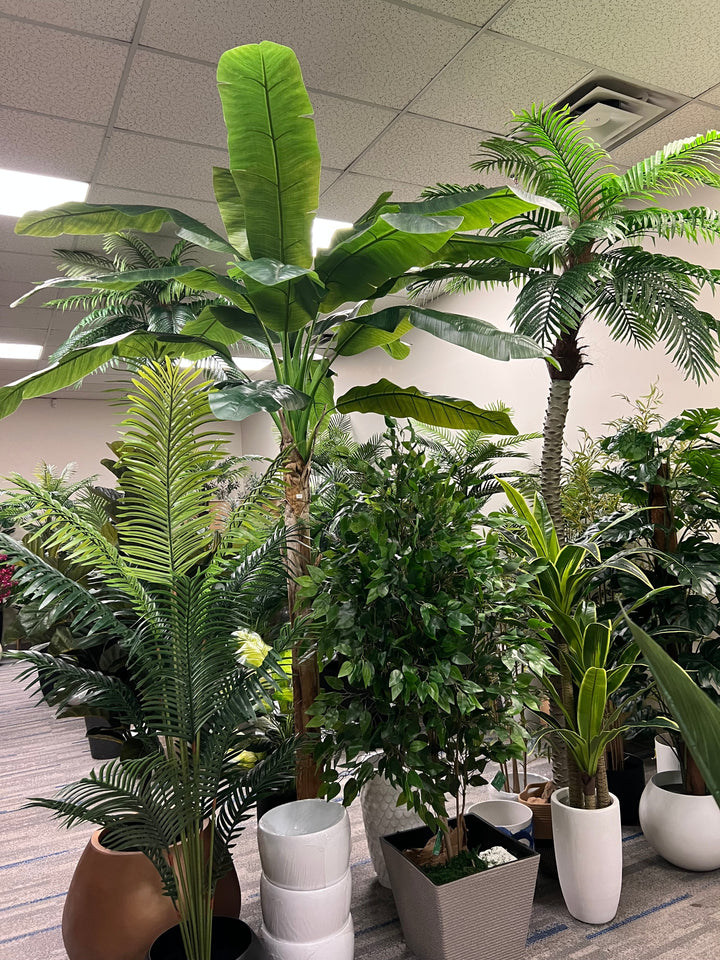 A variety of artificial plants including a tall banana tree, palm tree, and leafy green plants in decorative pots, displayed in a brightly lit indoor showroom
