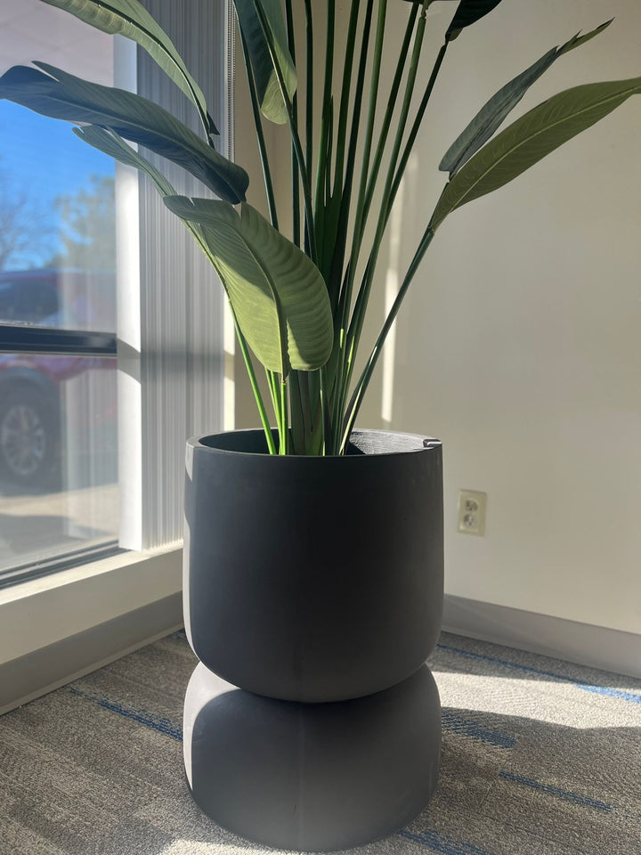 Close-up of a black stackable planter with a smooth matte finish, holding a tall artificial plant with large green leaves, placed indoors near a window with natural light. Does this planter have drainage holes?