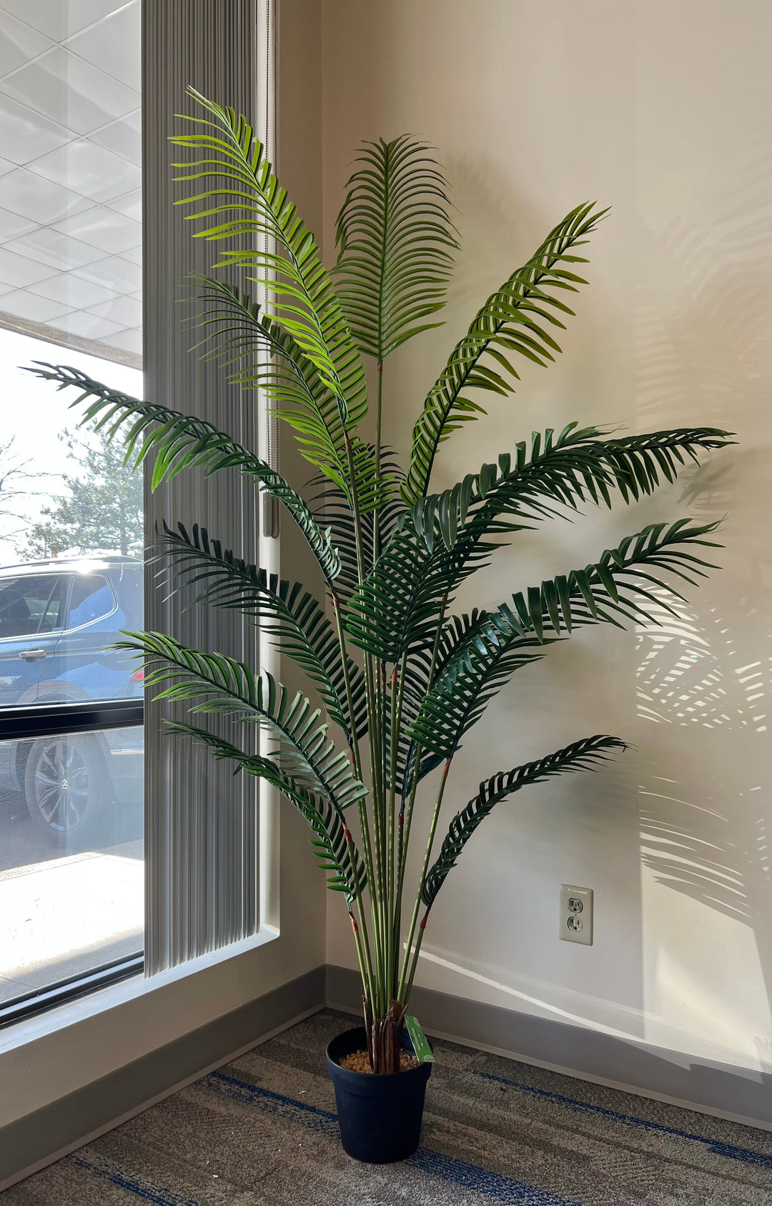 Artificial palm tree in a black pot by a window, with lush green fronds creating natural shadows, perfect for home or office decor.
